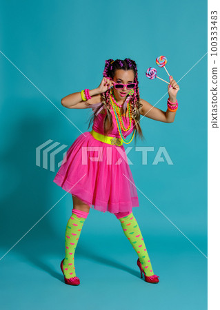 Lovely girl with a multi-colored braids hairstyle and bright make-up, posing in studio against a blue background, holding a lollipop in her hand. 100333483