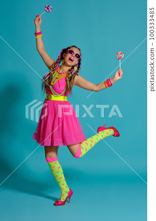 Lovely girl with a multi-colored braids hairstyle and bright make-up, posing in studio against a blue background, holding a lollipop in her hand. 100333485