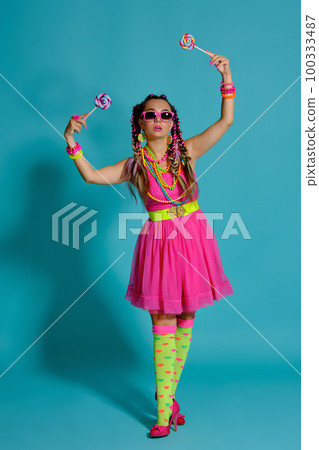 Lovely girl with a multi-colored braids hairstyle and bright make-up, posing in studio against a blue background, holding a lollipop in her hand. Lovely girl with a multi-colored braids hairstyle and bright make-up, posing in studio against a blue background, holding a lollipop in her hand. 100333487