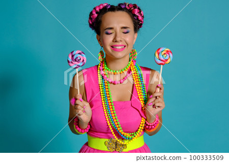 Lovely girl with a multi-colored braids hairstyle and bright make-up, posing in studio against a blue background, holding a lollipop in her hand. 100333509