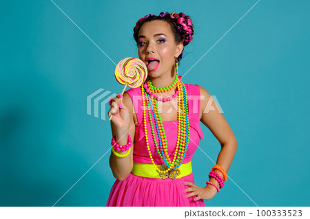 Lovely girl with a multi-colored braids hairstyle and bright make-up, posing in studio against a blue background, holding a lollipop in her hand. Lovely girl with a multi-colored braids hairstyle and bright make-up, posing in studio against a blue background, holding a lollipop in her hand. 100333523