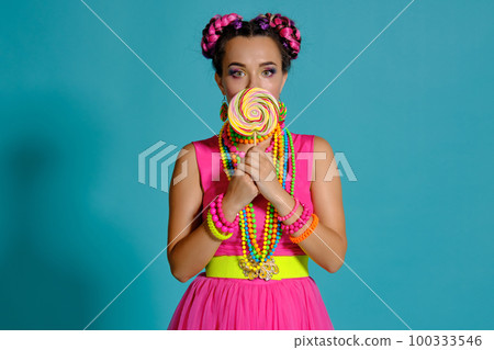 Lovely girl with a multi-colored braids hairstyle and bright make-up, posing in studio against a blue background, holding a lollipop in her hand. 100333546