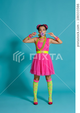 Lovely girl with a multi-colored braids hairstyle and bright make-up, posing in studio against a blue background, holding a lollipop in her hand. Lovely girl with a multi-colored braids hairstyle and bright make-up, posing in studio against a blue background, holding a lollipop in her hand. 100333566