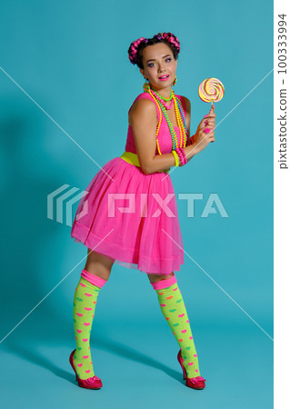 Lovely girl with a multi-colored braids hairstyle and bright make-up, posing in studio against a blue background, holding a lollipop in her hand. Lovely girl with a multi-colored braids hairstyle and bright make-up, posing in studio against a blue background, holding a lollipop in her hand. 100333994