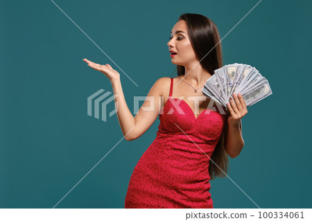 Brunette girl with a long hair, wearing a sexy red dress is posing holding a fan of hundred dollar bills against a blue background. Brunette girl with a long hair, wearing a sexy red dress is posing holding a fan of hundred dollar bills against a blue background. 100334061