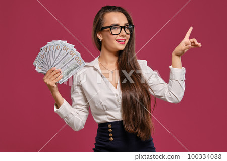 Brunette girl in glasses, wearing in a black short skirt and white blouse is posing holding a fan of hundred dollar bills against a pink background. Brunette girl in glasses, wearing in a black short skirt and white blouse is posing holding a fan of hundred dollar bills against a pink background. 100334088