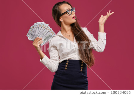 Brunette girl in glasses, wearing in a black short skirt and white blouse is posing holding a fan of hundred dollar bills against a pink background. 100334095