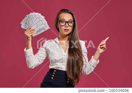 Brunette girl in glasses, wearing in a black short skirt and white blouse is posing holding a fan of hundred dollar bills against a pink background. 100334096