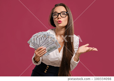 Brunette girl in glasses, wearing in a black short skirt and white blouse is posing holding a fan of hundred dollar bills against a pink background. Brunette girl in glasses, wearing in a black short skirt and white blouse is posing holding a fan of hundred dollar bills against a pink background. 100334098