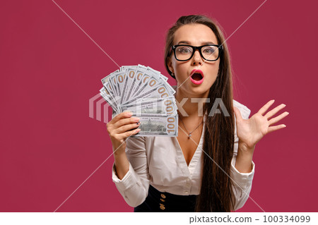 Brunette girl in glasses, wearing in a black short skirt and white blouse is posing holding a fan of hundred dollar bills against a pink background. 100334099