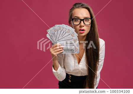 Brunette girl in glasses, wearing in a black short skirt and white blouse is posing holding a fan of hundred dollar bills against a pink background. 100334100