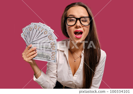 Brunette girl in glasses, wearing in a black short skirt and white blouse is posing holding a fan of hundred dollar bills against a pink background. 100334101