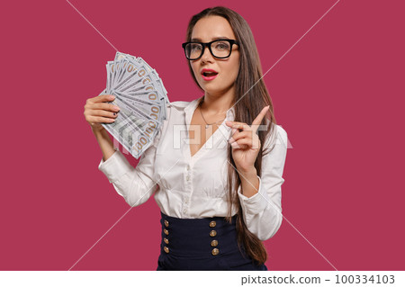 Brunette girl in glasses, wearing in a black short skirt and white blouse is posing holding a fan of hundred dollar bills against a pink background. 100334103
