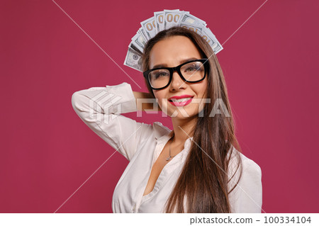 Brunette girl in glasses, wearing in a black short skirt and white blouse is posing holding a fan of hundred dollar bills against a pink background. Brunette girl in glasses, wearing in a black short skirt and white blouse is posing holding a fan of hundred dollar bills against a pink background. 100334104