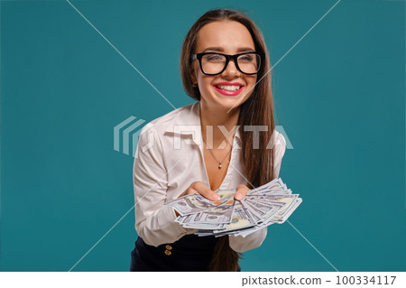 Brunette girl in glasses, wearing in a black short skirt and white blouse is posing holding a fan of hundred dollar bills against a blue background. Brunette girl in glasses, wearing in a black short skirt and white blouse is posing holding a fan of hundred dollar bills against a blue background. 100334117