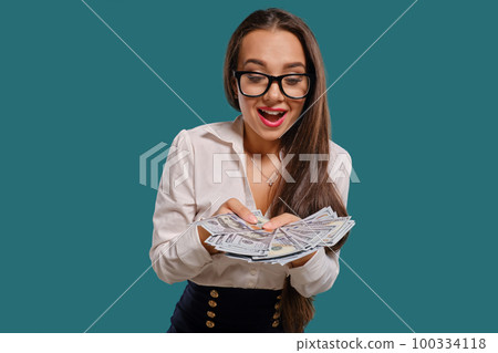 Brunette girl in glasses, wearing in a black short skirt and white blouse is posing holding a fan of hundred dollar bills against a blue background. Brunette girl in glasses, wearing in a black short skirt and white blouse is posing holding a fan of hundred dollar bills against a blue background. 100334118