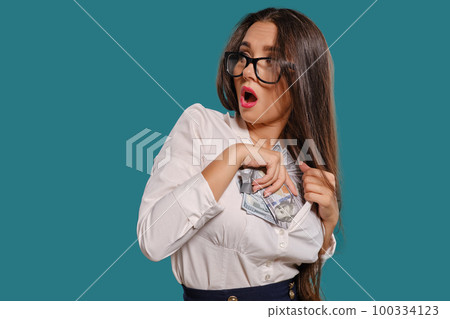 Brunette girl in glasses, wearing in a black short skirt and white blouse is posing holding a fan of hundred dollar bills against a blue background. Brunette girl in glasses, wearing in a black short skirt and white blouse is posing holding a fan of hundred dollar bills against a blue background. 100334123