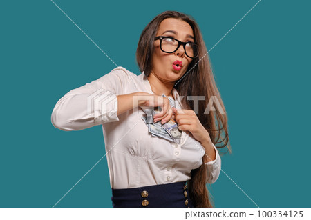 Brunette girl in glasses, wearing in a black short skirt and white blouse is posing holding a fan of hundred dollar bills against a blue background. Brunette girl in glasses, wearing in a black short skirt and white blouse is posing holding a fan of hundred dollar bills against a blue background. 100334125