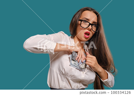 Brunette girl in glasses, wearing in a black short skirt and white blouse is posing holding a fan of hundred dollar bills against a blue background. Brunette girl in glasses, wearing in a black short skirt and white blouse is posing holding a fan of hundred dollar bills against a blue background. 100334126