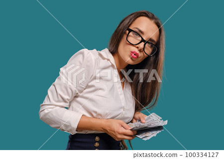 Brunette girl in glasses, wearing in a black short skirt and white blouse is posing holding a fan of hundred dollar bills against a blue background. Brunette girl in glasses, wearing in a black short skirt and white blouse is posing holding a fan of hundred dollar bills against a blue background. 100334127