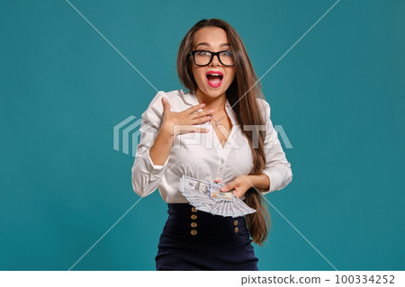 Brunette girl in glasses, wearing in a black short skirt and white blouse is posing holding a fan of hundred dollar bills against a blue background. 100334252