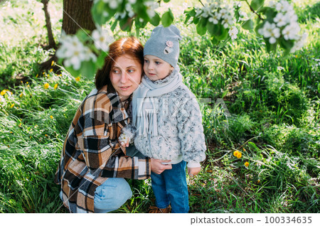 Young woman with her daughter in nature admires white flowering of spring trees Young woman with her daughter in nature admires white flowering of spring trees 100334635