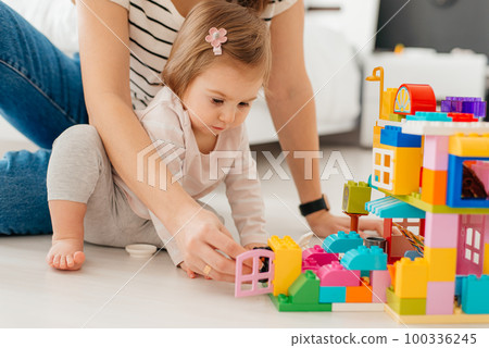 Laughing mother and little daughter playing colorful blocks, constructing tower, sitting on warm floor with underfloor heating, family enjoying leisure time together 100336245