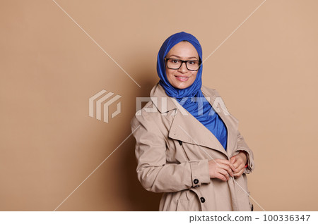 Gorgeous middle-aged confident t Middle-Eastern Muslim woman in beige coat and blue hijab, wearing stylish spectacles, smiling looking at camera, isolated on cream background. People Fashion Lifestyle Gorgeous middle-aged confident t Middle-Eastern Muslim woman in beige coat and blue hijab, wearing stylish spectacles, smiling looking at camera, isolated on cream background. People Fashion Lifestyle 100336347