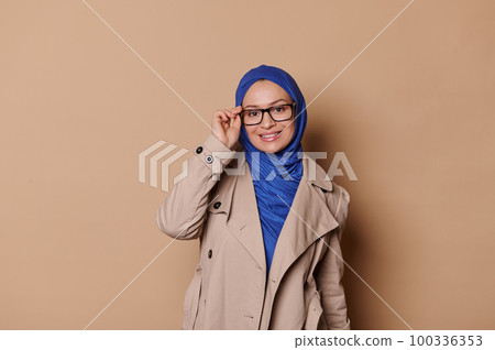 Attractive fashionable elegant Arab Muslim woman in blue hijab and casual beige coat, smiling looking at camera through stylish eyeglasses spectacles , isolated over cream background. People Fashion Attractive fashionable elegant Arab Muslim woman in blue hijab and casual beige coat, smiling looking at camera through stylish eyeglasses spectacles , isolated over cream background. People Fashion 100336353