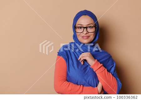 Confident portrait of a mature Muslim woman in blue hijab, wearing eyeglasses, smiling looking at camera, on isolated beige background. Islamic Middle-Eastern female fashion. Beautiful Arab women Confident portrait of a mature Muslim woman in blue hijab, wearing eyeglasses, smiling looking at camera, on isolated beige background. Islamic Middle-Eastern female fashion. Beautiful Arab women 100336362