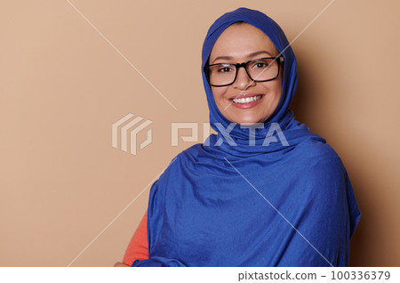 Confident middle-aged Arab Muslim woman with head covered in blue hijab, wearing stylish eyeglasses, smiling a toothy smile looking at camera, isolated on beige background with copy advertising space Confident middle-aged Arab Muslim woman with head covered in blue hijab, wearing stylish eyeglasses, smiling a toothy smile looking at camera, isolated on beige background with copy advertising space 100336379
