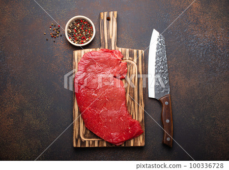 Raw uncooked top round beef steak on wooden cutting board with big kitchen knife and pepper on dark brown rustic stone background top view, cooking meat steak Raw uncooked top round beef steak on wooden cutting board with big kitchen knife and pepper on dark brown rustic stone background top view, cooking meat steak 100336728