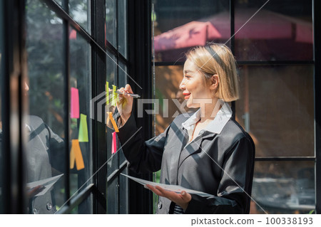 girl asian woman entrepreneur of small company putting a adhesive sticky notes in glass wall in office during analyzing formulating business strategies 100338193