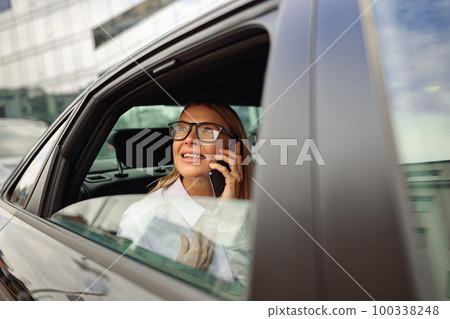 Businesswoman in eyeglasses traveling on back seat of car and talking phone while looking at window  100338248