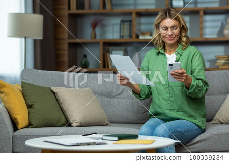 Work from home. An elderly woman in glasses is sitting on the sofa at home, holding a phone and documents in her hands. Works with papers, checks and pays online bills. Work from home. An elderly woman in glasses is sitting on the sofa at home, holding a phone and documents in her hands. Works with papers, checks and pays online bills. 100339284