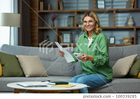 An elderly woman in glasses is sitting on the sofa at home, holding a phone and documents in her hands. Works with papers, checks and pays online bills. He looks at the camera, smiles. An elderly woman in glasses is sitting on the sofa at home, holding a phone and documents in her hands. Works with papers, checks and pays online bills. He looks at the camera, smiles. 100339285