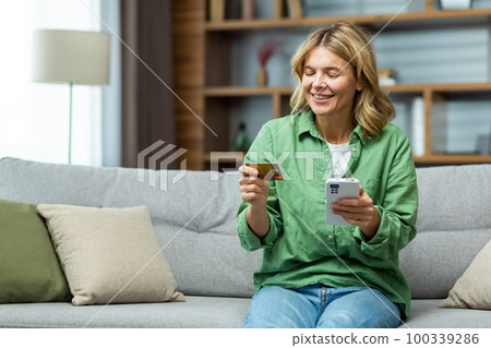 Smiling senior beautiful woman sitting on sofa at home. Holding a phone, using a credit card. Smiling makes online purchases, opened an account, checks. Smiling senior beautiful woman sitting on sofa at home. Holding a phone, using a credit card. Smiling makes online purchases, opened an account, checks. 100339286