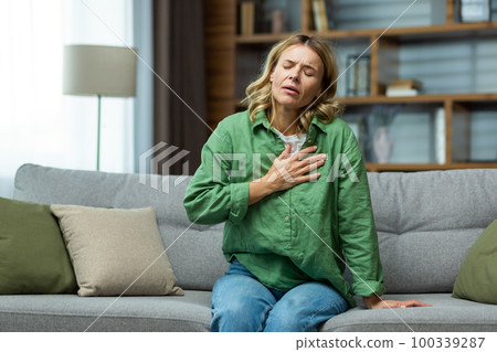 Heart attack, panic attack,. An elderly woman sits on the sofa at home and holds her hand to her chest. It is difficult to breathe, feels severe pain. Heart attack, panic attack,. An elderly woman sits on the sofa at home and holds her hand to her chest. It is difficult to breathe, feels severe pain. 100339287