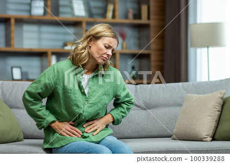 Unhealthy senior woman sitting on sofa at home. holding her stomach, grimacing in pain. She experiences severe abdominal pain, eating disorder, diarrhea, poisoning, menstruation, menopause. Unhealthy senior woman sitting on sofa at home. holding her stomach, grimacing in pain. She experiences severe abdominal pain, eating disorder, diarrhea, poisoning, menstruation, menopause. 100339288