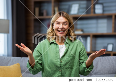 Close-up photo. Portrait of a young happy senior blonde woman sitting on the sofa at home wearing a green shirt. He smiles at the camera, enthusiastically spreads his hands, shows. Close-up photo. Portrait of a young happy senior blonde woman sitting on the sofa at home wearing a green shirt. He smiles at the camera, enthusiastically spreads his hands, shows. 100339289