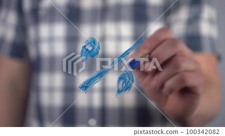 A young man draws a mathematical percent sign on the glass. A man in a shirt draws with a brush and blue paint. Close-up. 100342082