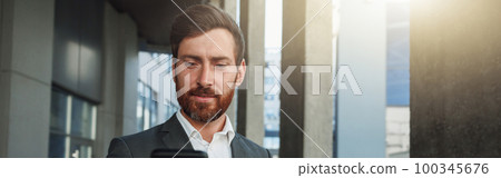 Attractive businessman holding phone standing near office building during break Attractive businessman holding phone standing near office building during break 100345676