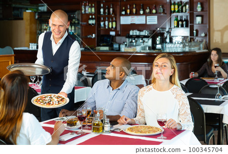 Polite waiter serving pizza to friendly company in pizzeria 100345704