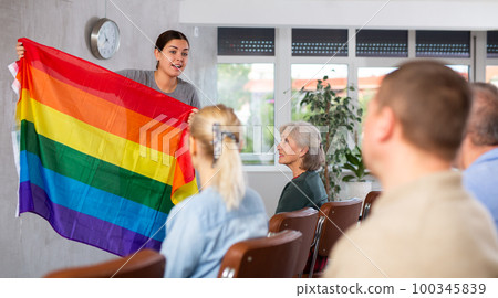 Portrait of young female teacher conducting lesson for adult students, talking about LGBT community and showing rainbow flag 100345839