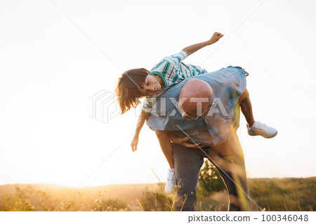 Photo of man carrying his grandson piggyback in the field. Boy and grandfather spending time together at summer. Brunette boy boy wearing striped t-shirt and man blue shirt and glasses. 100346048