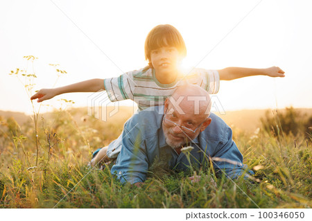 Little boy and his grandfather laying on a grass. Boy and grandfather spending time together at summer. Brunette boy wearing striped t-shirt and man blue shirt and glasses. 100346050