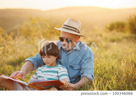 Little boy and his grandfather sitting on a grass and reading a book in the field. Boy and grandfather spending time together at summer. Brunette boy wearing striped t-shirt and man blue shirt and 100346066