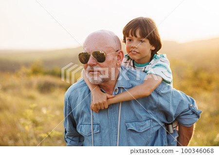 Photo of man carrying his grandson piggyback in the field. Boy and grandfather spending time together at summer. Brunette boy boy wearing striped t-shirt and man blue shirt and sunglasses. 100346068
