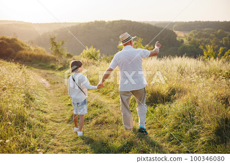 Backside photo of little boy and his grandfather walking in the field. Boy and grandfather spending time together at summer. Brunette boy and man wearing striped shirts. 100346080