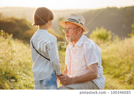 Photo of little boy and his grandfather standing and talking in the field. Boy and grandfather spending time together at summer. Brunette boy and man wearing striped shirts. 100346085